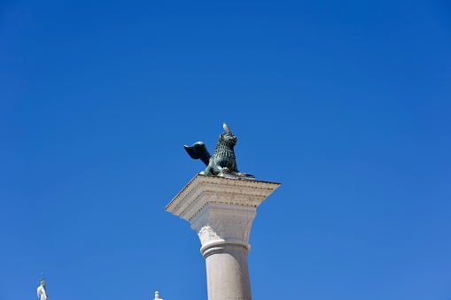 Bronze sculpture of lion with wings and book on top of stone column on a sunny summer day with seagull on his head at Italian City of Venice. Photo taken August 7th, 2023, Venice, Italy.