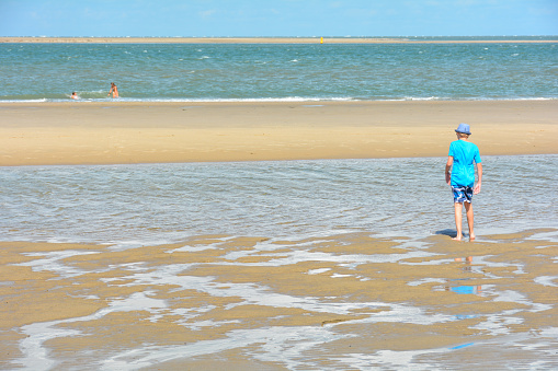 A boy in a hat runs on the sandy beach at low tide, with the sea and blue sky