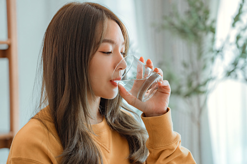 Asian woman drink water start new day with healthy life habit, Female holding glass drinking clean mineral natural still water in living room at home morning, Lifestyle healthcare concept