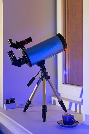 A modern telescope stands on the windowsill and is pointed out the window. Blue catadioptric astronomical telescope on a tripod in the room