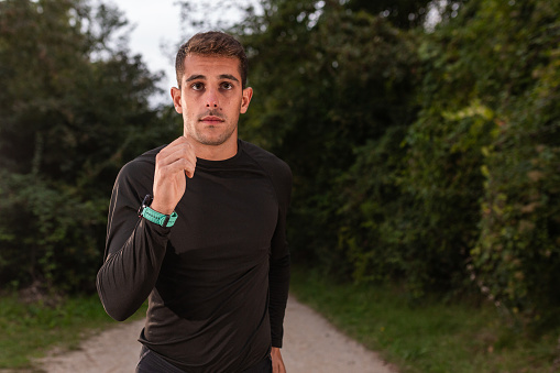 a solitary figure standing on a dirt path, surrounded by lush greenery. The individual, dressed in a black long-sleeved shirt, appears to be pausing during a walk or run.