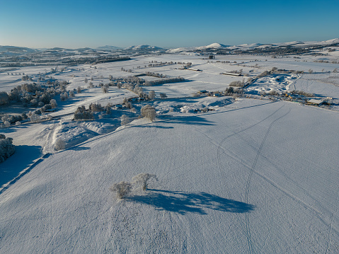 Aerial drone view of a snow covered winter landscape, on a sunny morning in the Scottish Borders. The tranquil scene looks across agricultural fields and open countryside, with hills in the far distance.