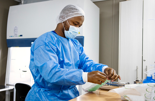 Male pharmacist in protective mask and smock opening a packet of gloves at a table in a medical lab