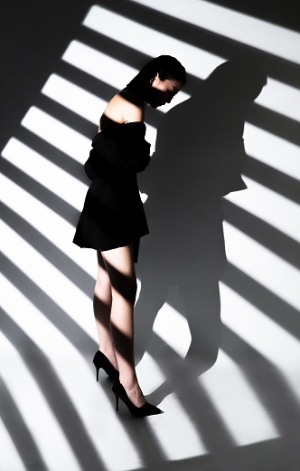 Fashion model posing in black dress and black high heels in front of white background with striped shadows