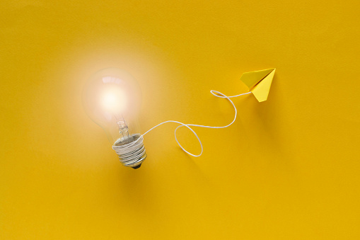 Bright light bulb attached to a flying  yellow paper plane with a string  on yellow background.