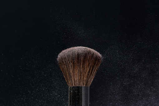 Make-up brush with beige nude color powder splashes explosion on black background. Beauty concept.