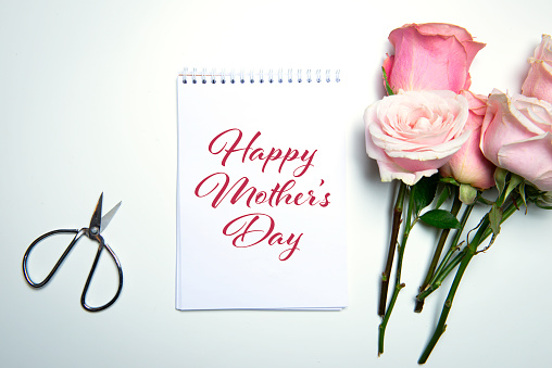 Scissors, notepad with Happy Mother’s Day text and roses on white table.