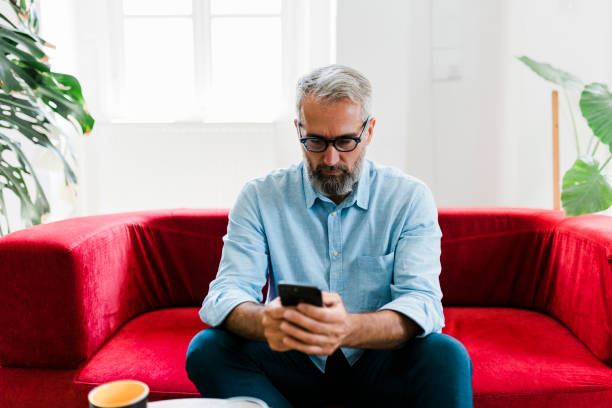 istock Senior man using smart phone and sitting on sofa at living room 1955632787