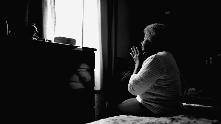 One tranquil senior African American lady in 80s praying at home with eyes closed. One Hopeful and grateful black woman in PRAYER in monochromatic