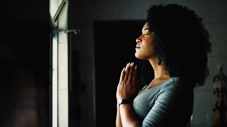 One Spiritual young black woman standing by window at home in prayer with eyes closed. African American adult girl with curly hair engaged in deep meditation, profile face and hands clenched together