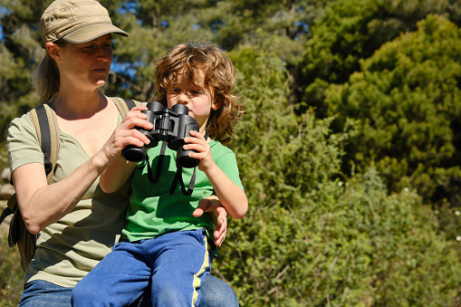 Young mother in casual clothes and cap with little son sitting in green forest with binoculars while enjoying sunny day together against blurred trees background