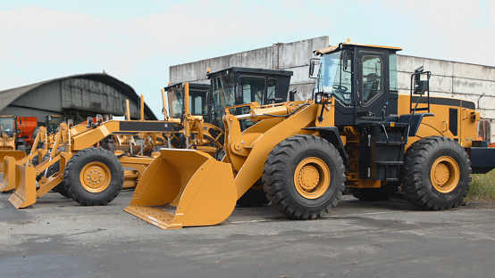 Yellow wheel loaders and graders stand in a row near old hangars