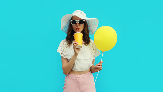 Portrait of beautiful caucasian young woman model with cup of coffee or fresh juice holding yellow balloon wearing white summer straw hat on studio blue background