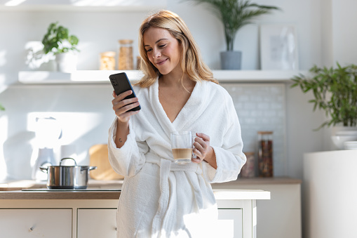 Shot of smiling young woman enjoying a cup of coffee while using her mobile phone in the living room at home.