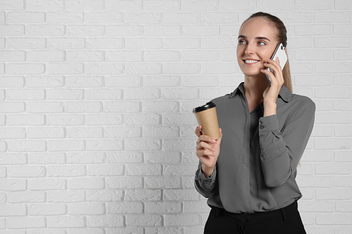 Happy young secretary with paper cup of drink talking on smartphone near white brick wall, space for text