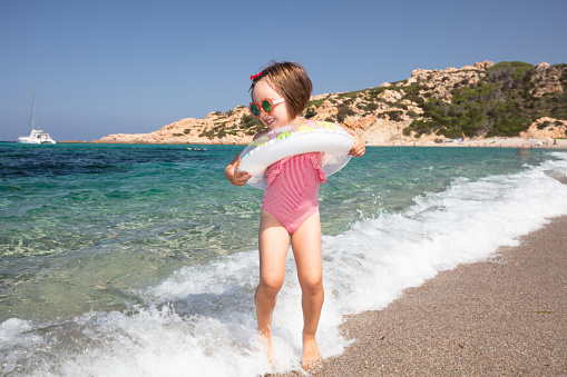 A little girl wearing a colorful lifebuoy bathes in the beautiful and colorful waters of the Mediterranean Sea on a summer day on the beach of Monti Russu in Santa Teresa Gallura, facing the Strait of Bonifacio.