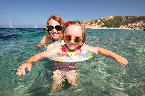 A little girl is playing with her mother in the beautiful and colorful waters of the Mediterranean Sea on a summer day on the beach of Monti Russu in Santa Teresa Gallura, facing the Strait of Bonifacio.