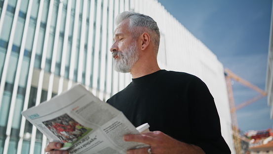 Successful senior reading newspaper on street closeup. Handsome grey bearded man holding coffee cup enjoying morning news at downtown buildings. Brutal ceo investor looking camera outdoors