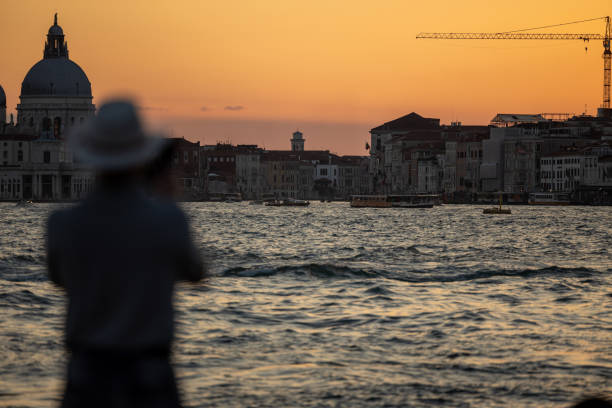 A beautifully atmospheric scene of a gondolier navigating the calm waters of Venice at twilight. The soft evening light casts a warm glow on the distant cityscape, while the wooden mooring poles in the foreground add depth to the composition. The blurred motion of the gondolier creates a dreamy and romantic ambiance, perfectly capturing the timeless charm of Venetian waterways.