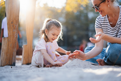 Cute toddler girl playing in sand on outdoor playground. Beautiful baby having fun on sunny warm summer sunny day. Happy healthy child with sand toys and in colorful fashion clothes