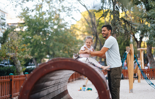 A side view of a smiling Caucasian man and his pretty baby girl looking away while having fun in the park.