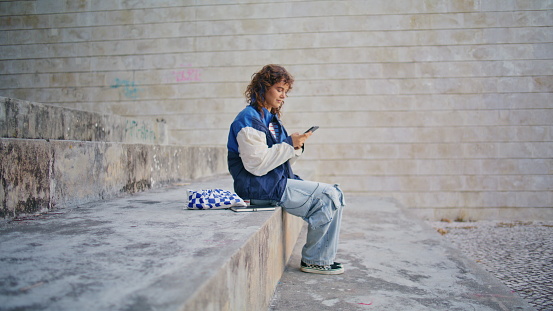 Calm girl watching cell phone screen on street wearing trendy casual outfit. Curly carefree woman putting smartphone on concrete step relaxing outdoors. Stylish informal hipster sitting urban district