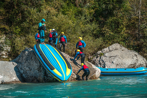 The thrill of river rafting takes a playful turn as a woman in a yellow helmet slides down an upturned raft, her teammates cheering her on in a moment of pure fun.