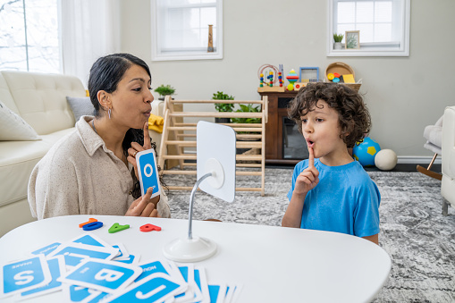 A female speech therapist works with a young boy as they practice their sounds and letter enunciation.  The Therapist is dressed casually and is holding up alphabetical cue cards as they work together in front of a mirror.