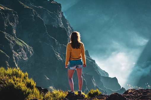 Description: Sporty female toursit overlooks the breathtaking depths of a cloud-covered valley and enjoys the view of the volcanic mountain landscape. Pico do Arieiro, Madeira Island, Portugal, Europe.
