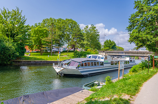 Amiens, France, July 3, 2023: motor ship tourist boat is sailing down narrow Somme river water with car bridge behind and green trees on riverbank shore, Hauts-de-France Region, Northern France