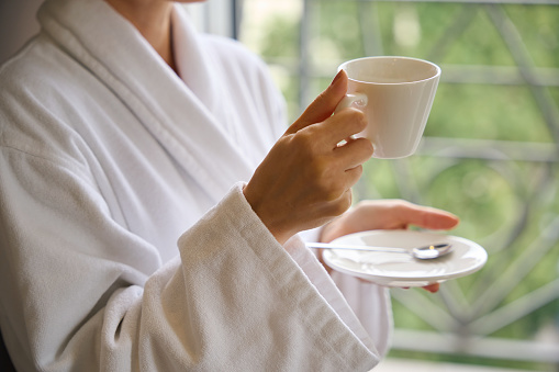 Cropped photo of lady dressed in bathrobe holding ceramic saucer and cup in her hands