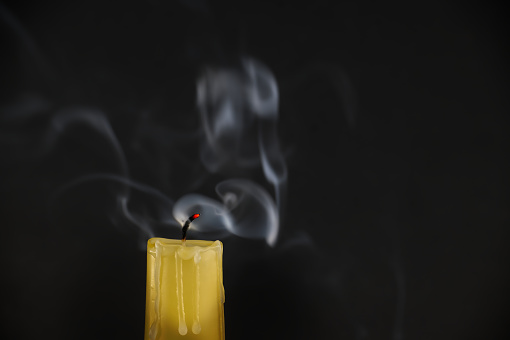 Yellow candle and its wick, which appears red is still emitting smoke after the flame has been extinguished. White smoke is rising in various directions, forming delicate pattern against dark backdrop