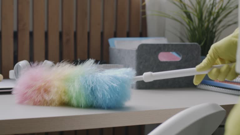 Close-up of colorful duster wiping table surface while person in rubber gloves dusting furniture in apartment