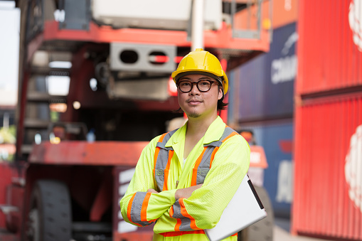 Portrait of Asian male container yard worker standing with crossed arms at commercial dock site. Foreman or supervisor inspecting container boxes from cargo freight ship