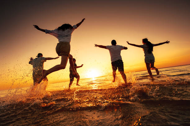 Group of happy young friends run and jump at sunset beach Silhouettes of big group of happy young friends are having fun, run and jump at calm sunset lake beach teens interacting photos stock pictures, royalty-free photos & images