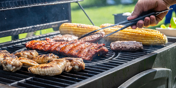 Pork ribs, chicken wings and corn being roasted on grill and flipped with tongs at barbecue party close up