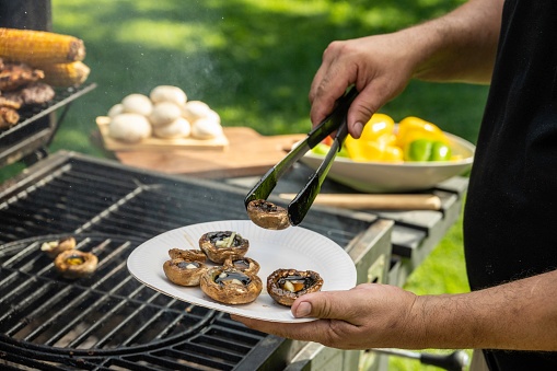 Man holding tongs and putting grilled mushrooms on plate outdoors at barbecue party