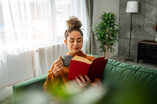 The photo shows a young beautiful woman in her comfortable apartment indulging in her favorite book