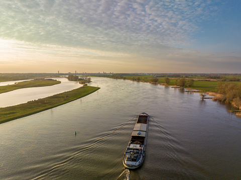 Panoramic aerial view on a freight ship sailiing on the river IJssel during a springtime sunset in Overijssel. The flow of the river is leading towards the setting sun in the distance while lights are popping up in the city at the end of a beautiful springtime day.