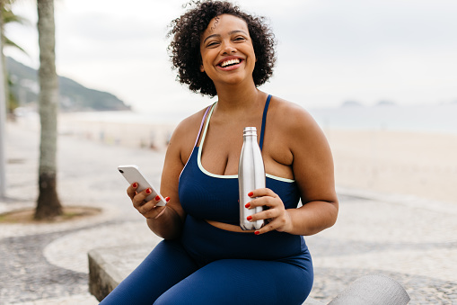 Fit woman in sportswear sitting on a beach promenade, holding a smartphone and enjoying a workout break. Happy young woman browsing the best beach workouts as she engages in a healthy lifestyle.