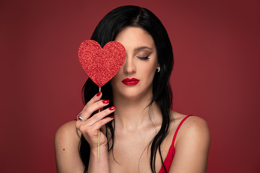 Beauty studio portrait of young beautiful woman holding red heart against red background