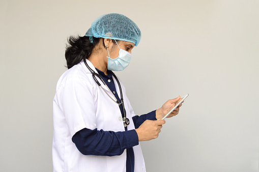 Side view One Indian Asian woman or lady or female doctor wearing surgical mask, cap, and white lab coat and a stethoscope around her neck holding an ipad or tablet in hands over grey horizontal  background with copy space for text
