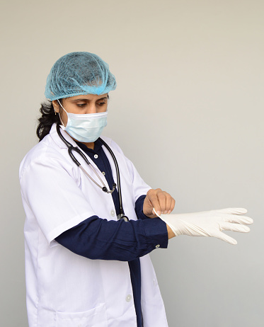 Vertical photograph of one female surgeon with blue shirt, white lab coat, stethoscope getting ready by putting on surgical gloves in her hand over grey background with copy space for text.