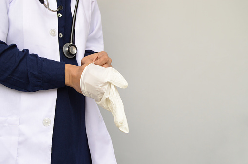 Horizontal photograph of one unidentifiable surgeon with blue shirt, white lab coat, stethoscope getting ready by putting on surgical gloves in her hand over grey background with copy space for text. The person is at the left side leaving right side free for text. Just the body is visible while the face is not. The gear is for protection from coronavirus or other infections.