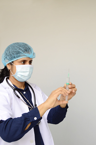 Vertical photo of a woman nurse or doctor or health care worker, physician injecting vaccine with syringe in her hand to administer a dose over gray background with copy space for text.