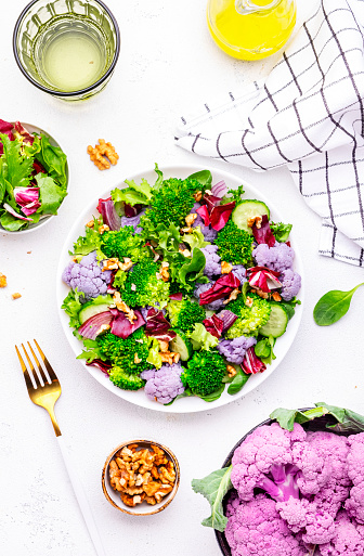 Yummy vegan salad with purple cauliflower, broccoli, fresh cucumbers, onion, lettuce with walnuts, white table background, top view