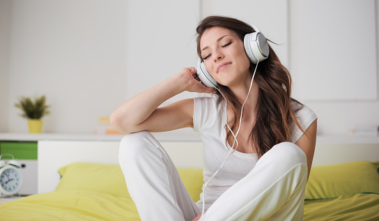 Cheerful girl with headphones relaxing at home, she is sitting on the bed and listening to her favourite music