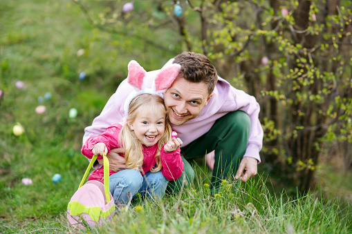 Caucasian father and daughter with bunny ears  collecting Easter eggs  and putting Easter eggs in baskets in the park. Easter egg hunt concept