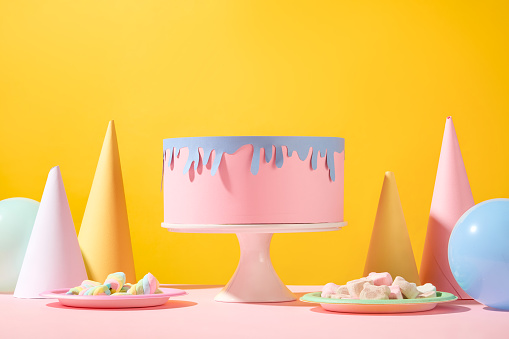 A birthday cake folded from colored paper is displayed on a pedestal, plates of marshmallows, birthday cones and balloons on a pink-yellow background.