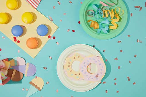 Macarons, ribbons, ice cream and donuts are cut from colored paper, confetti is decorated on a blue background. Colorful layout with birthday theme.